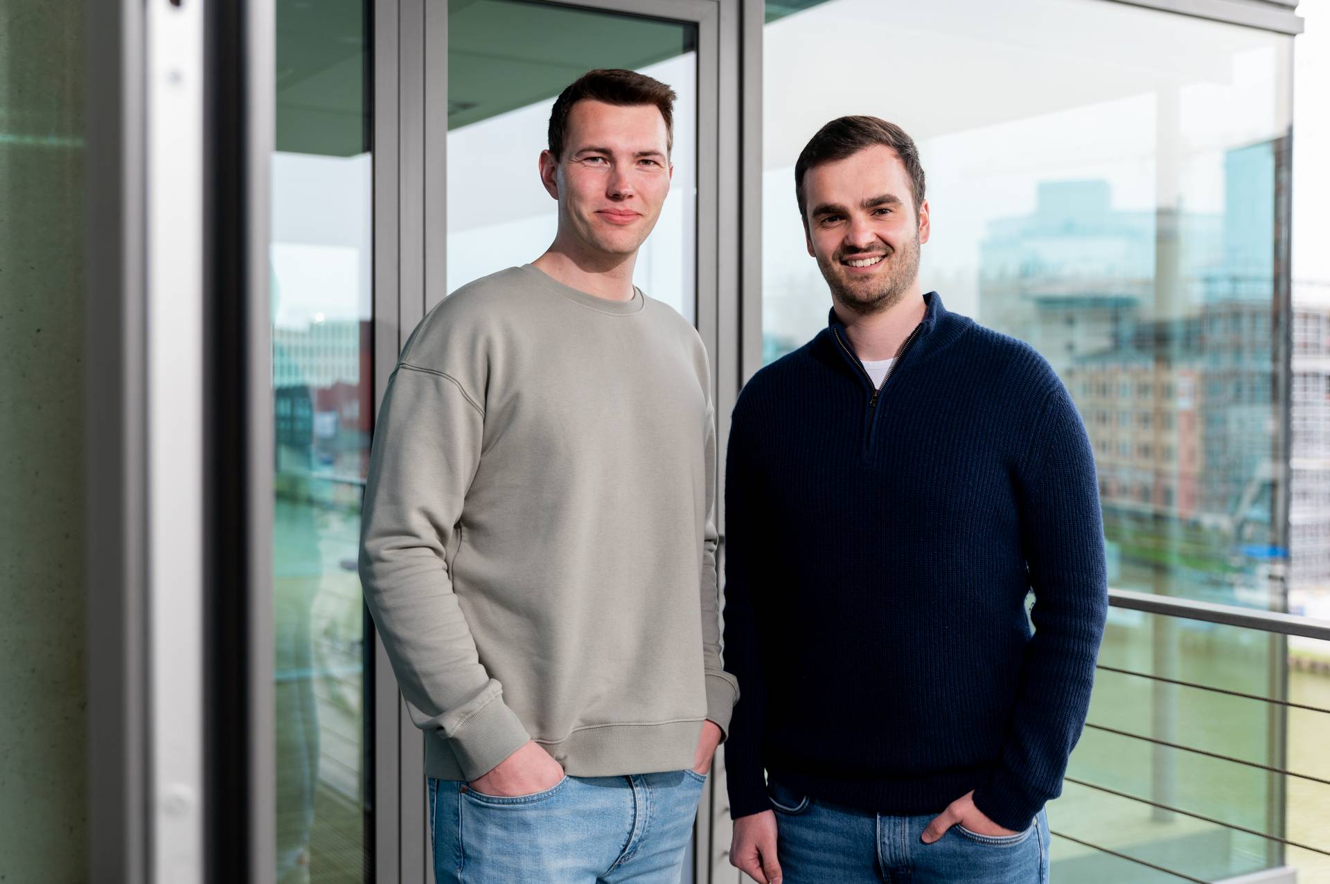 Symbolbild "Umsatzsteuer ID": Jan Philipp und Tobias, Steuerberater der Hansel & Vogt Steuerberatung, blicken in die Kamera und stehen vor einer Glasfassade. Tobias trägt ein beiges Sweatshirt, Jan Philipp einen dunkelblauen Pullover.